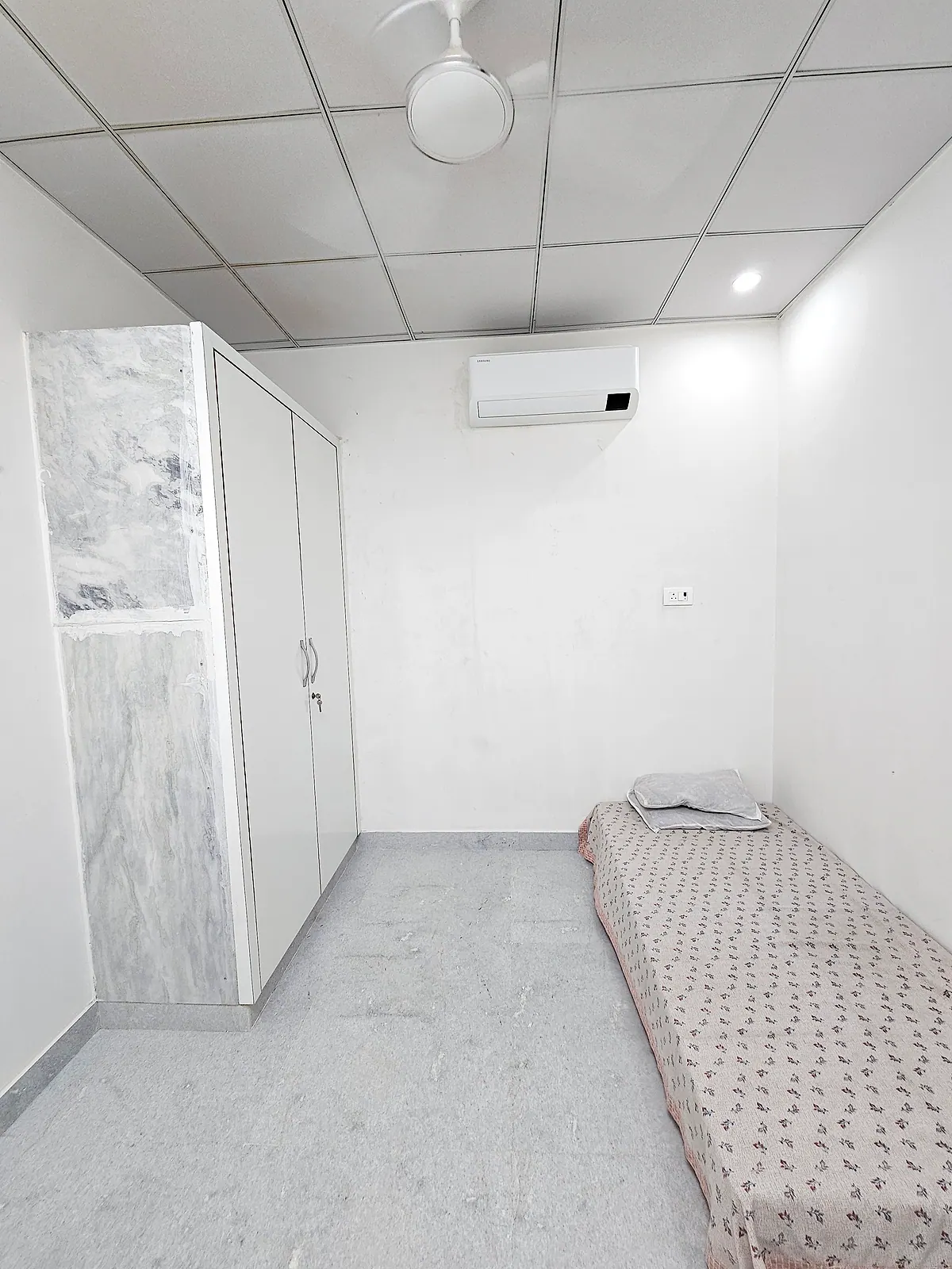 Wider view of the representative furnished room showing the bed, wardrobe, desk, chair, AC and ceiling fan